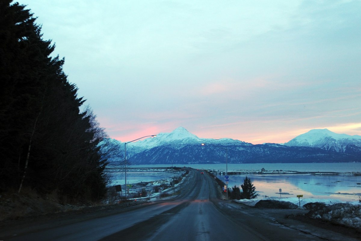 A two-lane road curving toward snow-covered mountains on the Kenai Peninsula near Homer