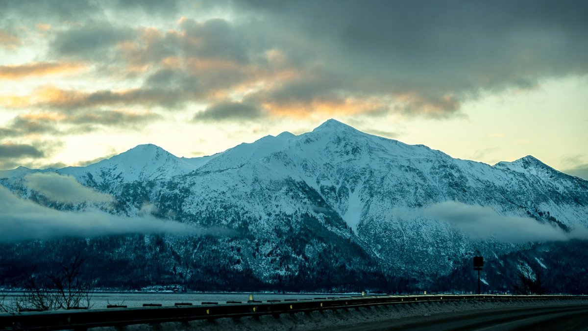 Snow-covered mountains rising above a cloudy Alaskan sky along a scenic highway