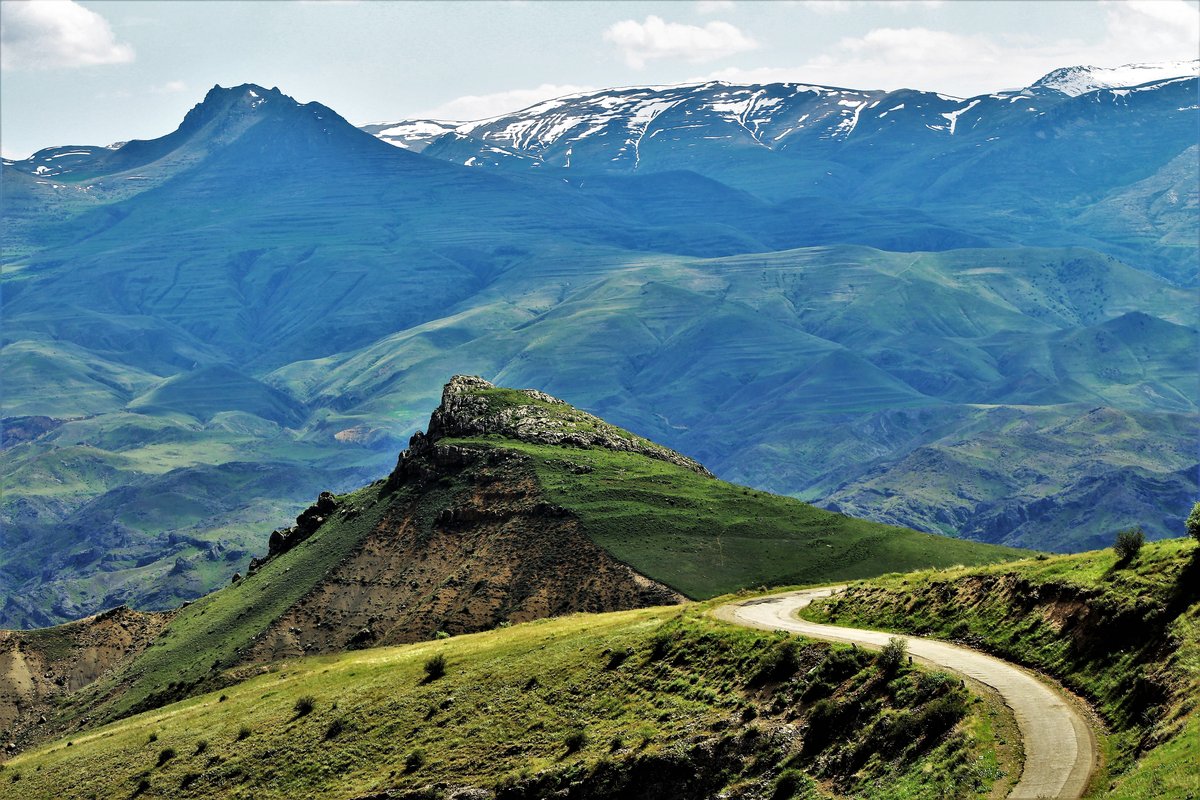 A winding mountain road through green hills in Vayots Dzor, with a rocky ridge in the foreground and snow-streaked peaks beyond