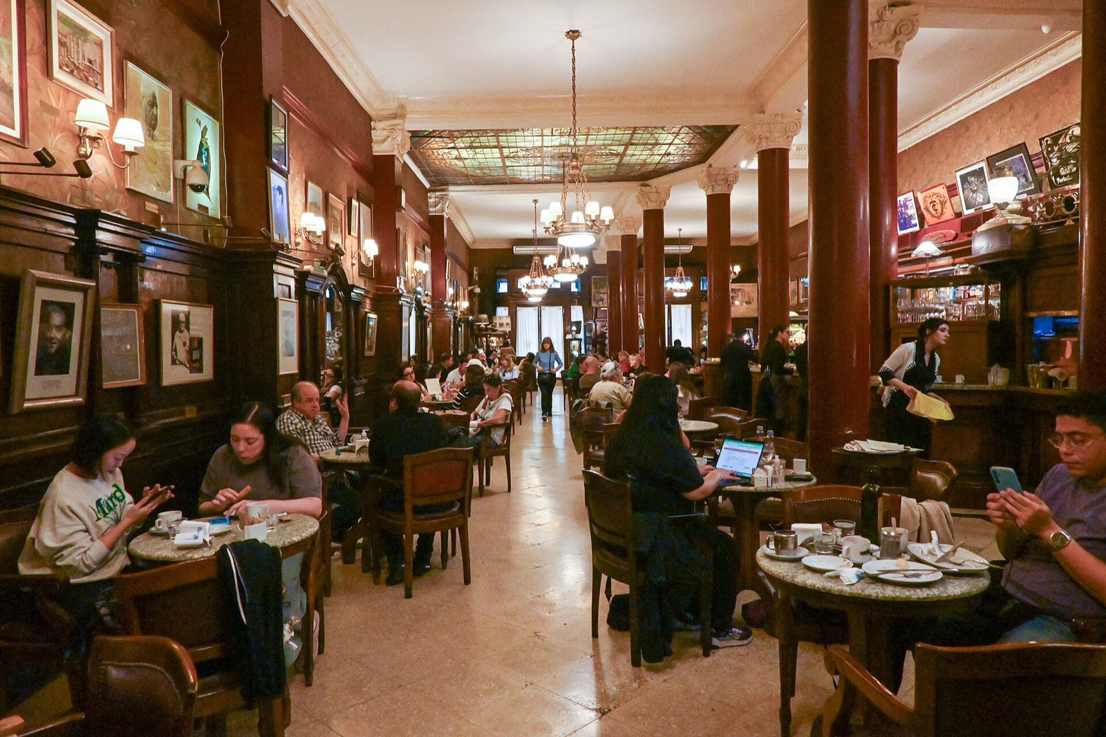 Busy interior of Café Tortoni with stained-glass skylight, ornate columns, chandeliers, and marble tables