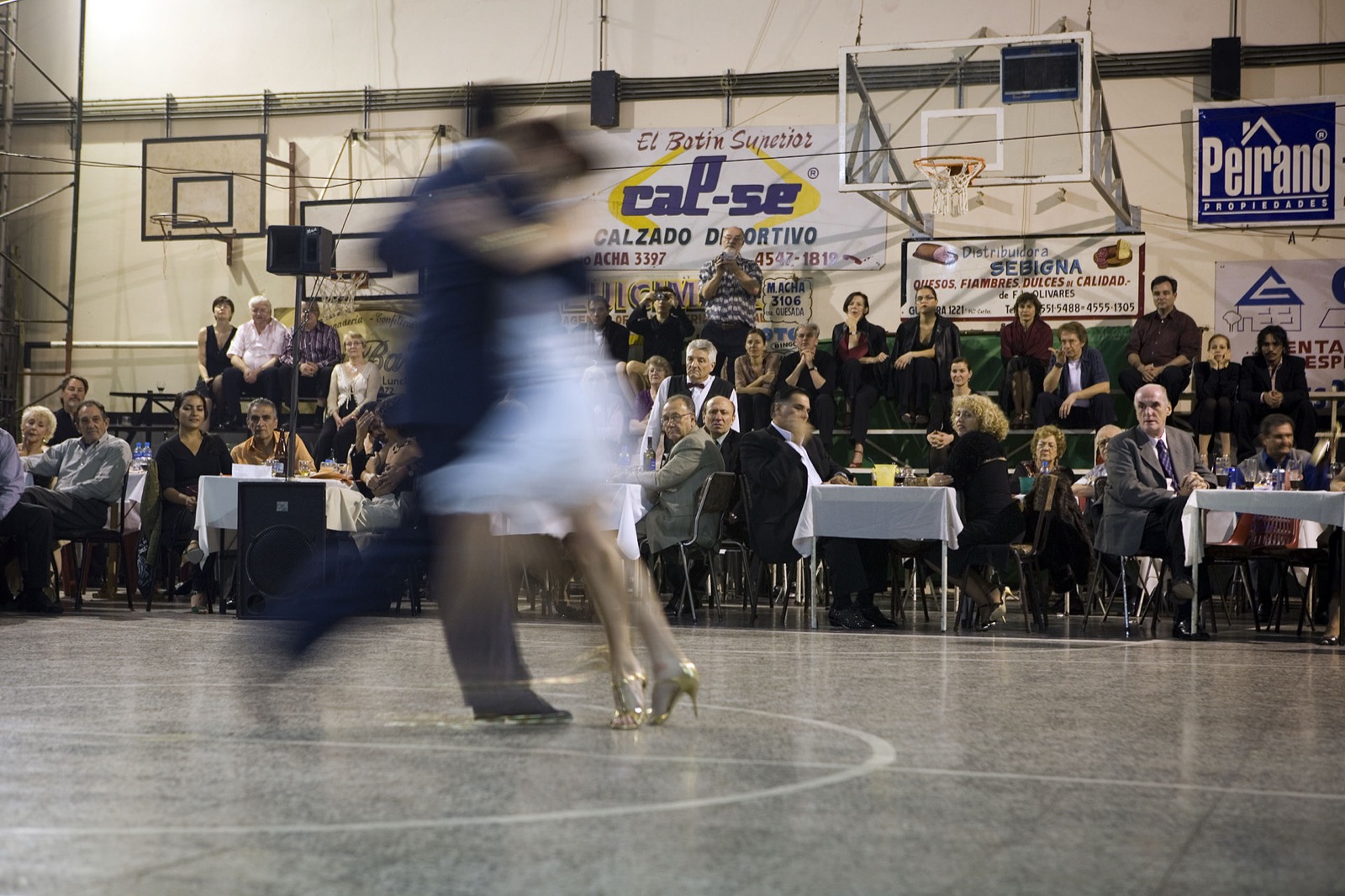 A couple dancing tango on the floor of a milonga in Buenos Aires while spectators watch from tables and bleachers