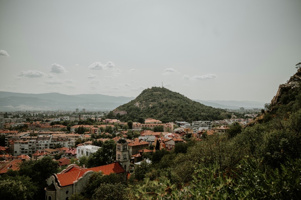 Panoramic view of Plovdiv from a hillside, with a forested tepe, red-tiled rooftops and mountains beyond