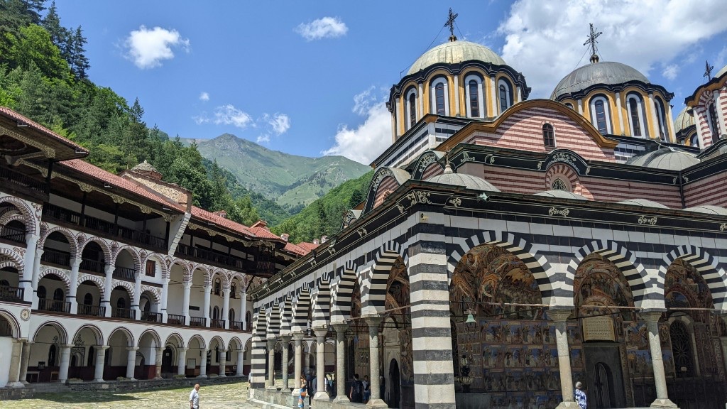 Rila Monastery courtyard with black-and-white striped church arcades, frescoed portico and forested mountain slopes beyond