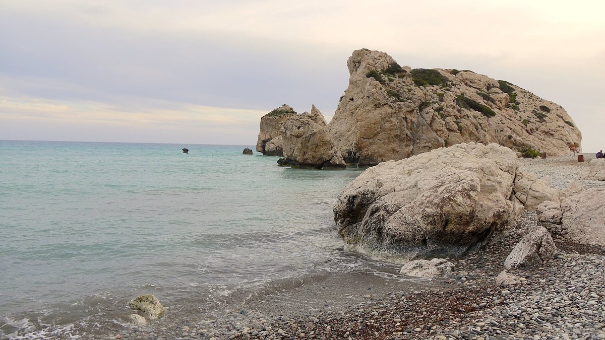 Aphrodite's Rock, a large limestone sea stack rising from calm water beside a pebble beach near Paphos