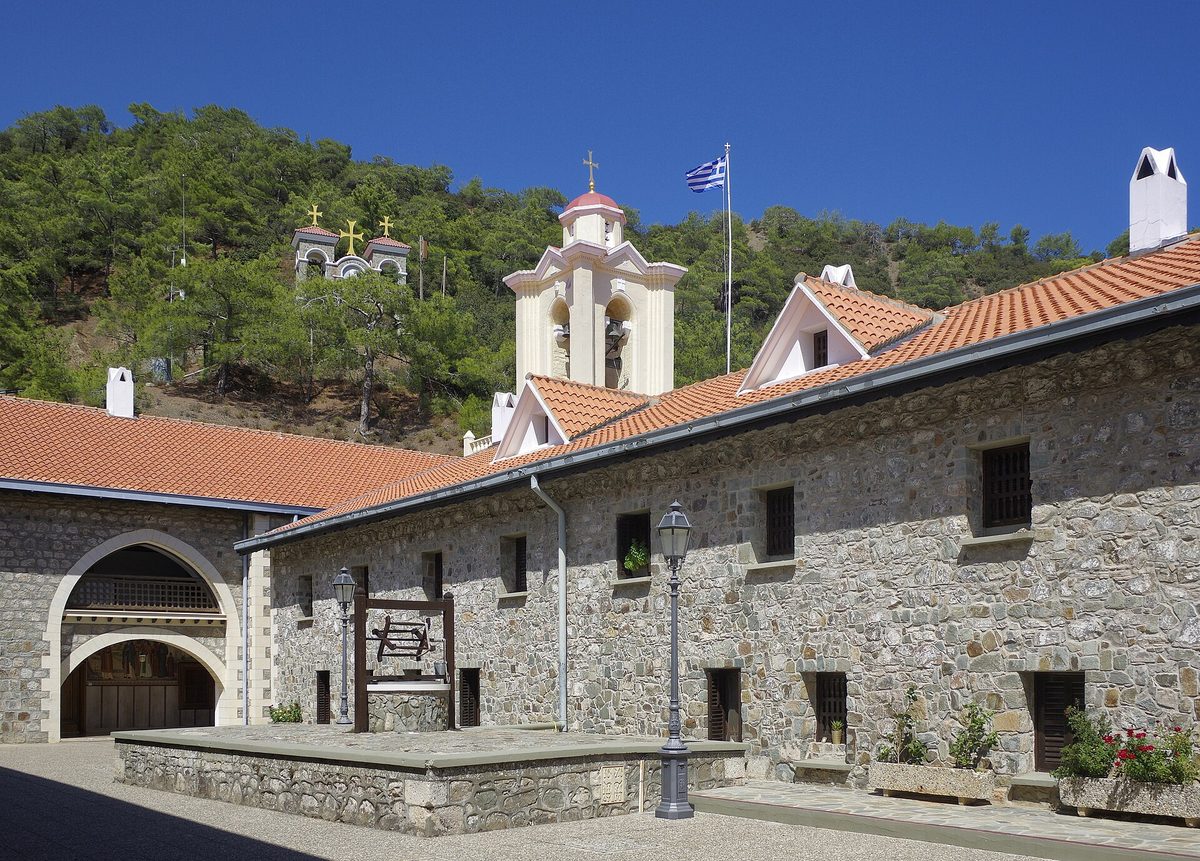 Stone buildings and bell tower of Kykkos Monastery with red-tiled roofs against a forested hillside