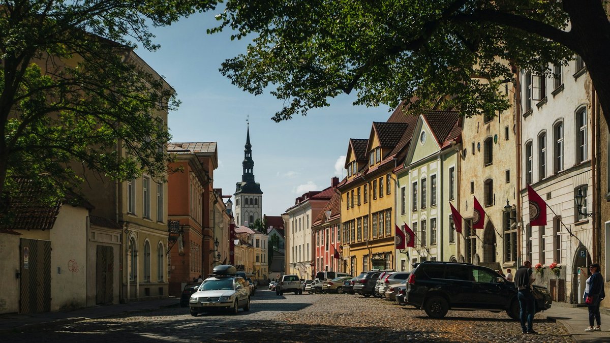Calle empedrada en la Ciudad Vieja de Tallin, coches aparcados y aguja de iglesia al fondo