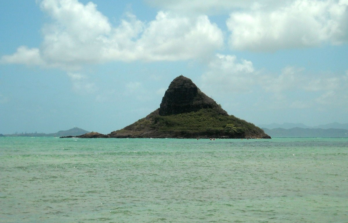 A small green island in turquoise water off Oahu, with the Koʻolau mountains behind it under a cloudy sky