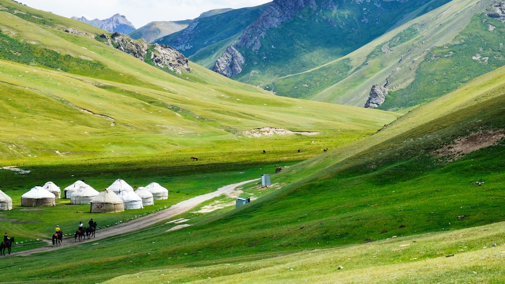 Traditionelle weiße Jurten auf üppigen grünen Almwiesen unter schroffen Berggipfeln in Kirgisistan, mit Reitern auf einem Schotterweg durch die Hochgebirgslandschaft