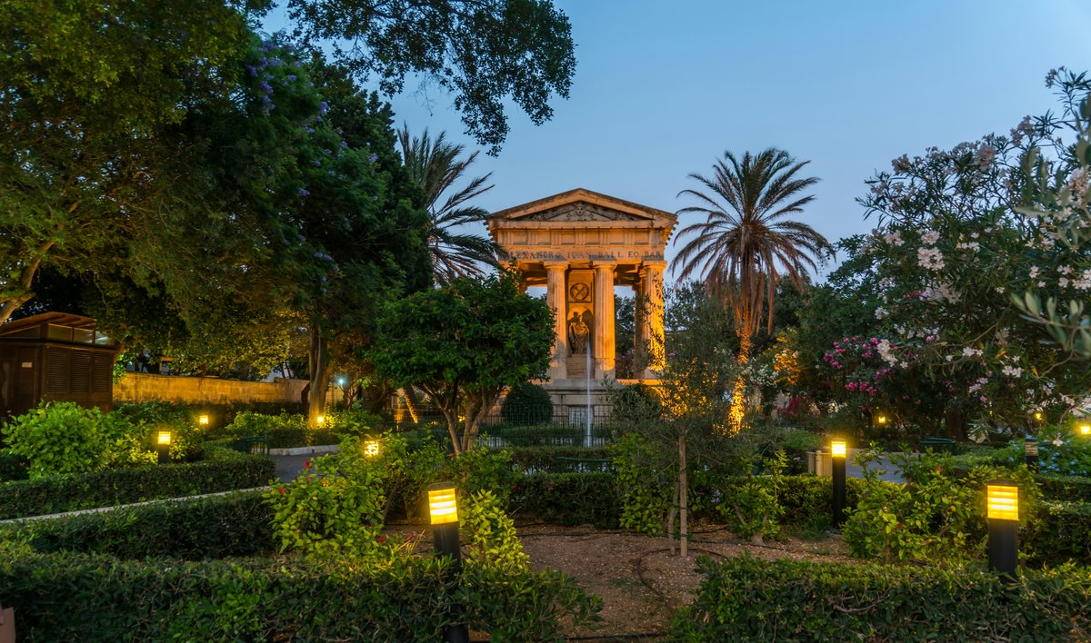 The Monument to Sir Alexander Ball lit up at dusk in the Lower Barrakka Gardens, Valletta, surrounded by palm trees and hedges