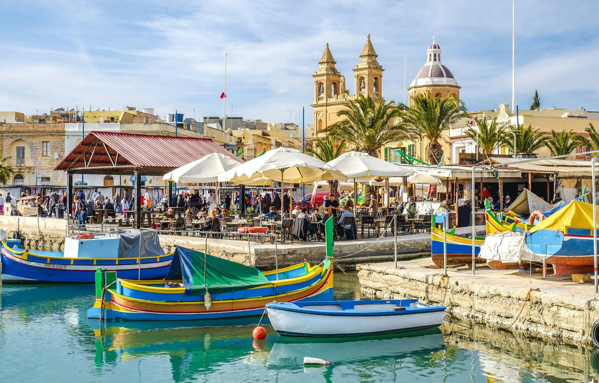 Colourful luzzu fishing boats moored along the Marsaxlokk waterfront beside market stalls and the parish church