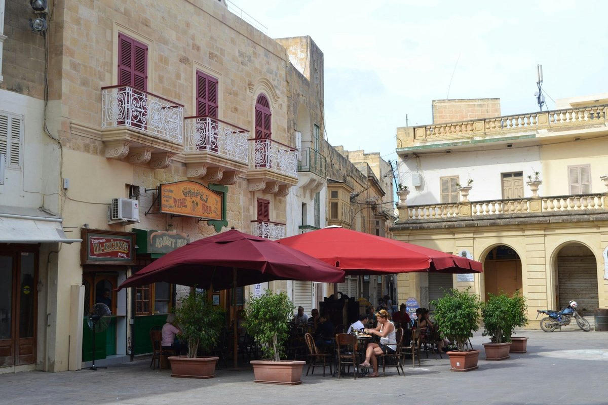 Outdoor café tables under red umbrellas in St. George's Square, Victoria, Gozo, framed by limestone buildings with balconies
