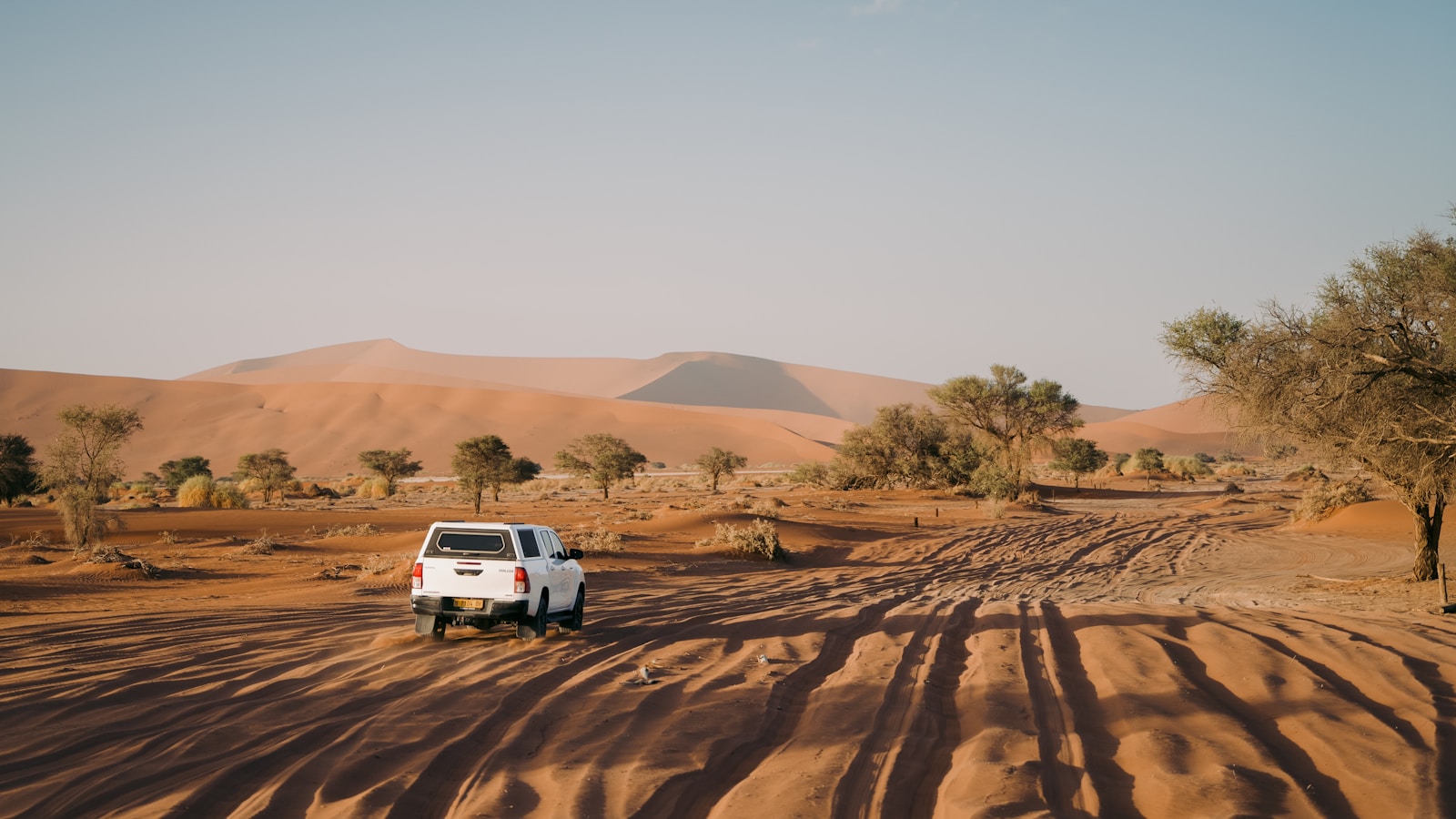 White pickup truck driving on a sandy desert road toward orange dunes in Namibia