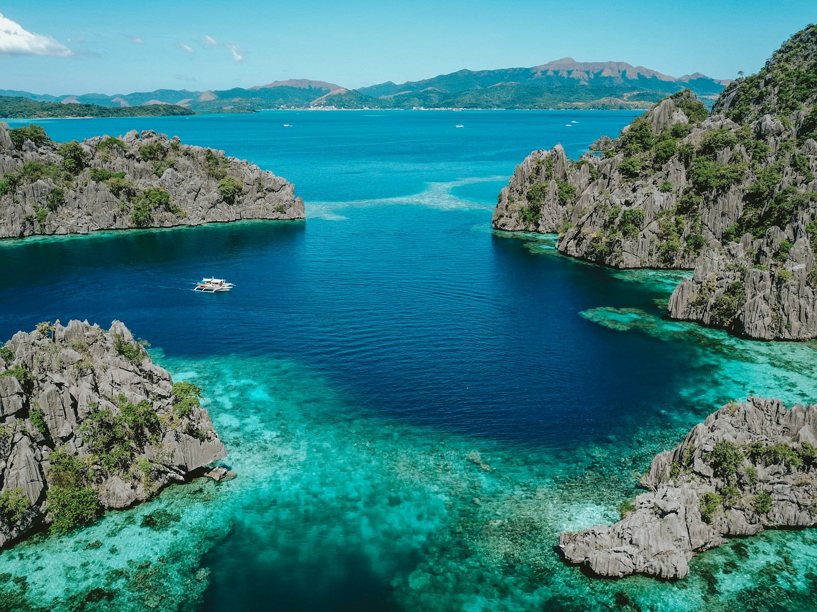Aerial view of a clear blue lagoon surrounded by limestone mountains with a small outrigger boat