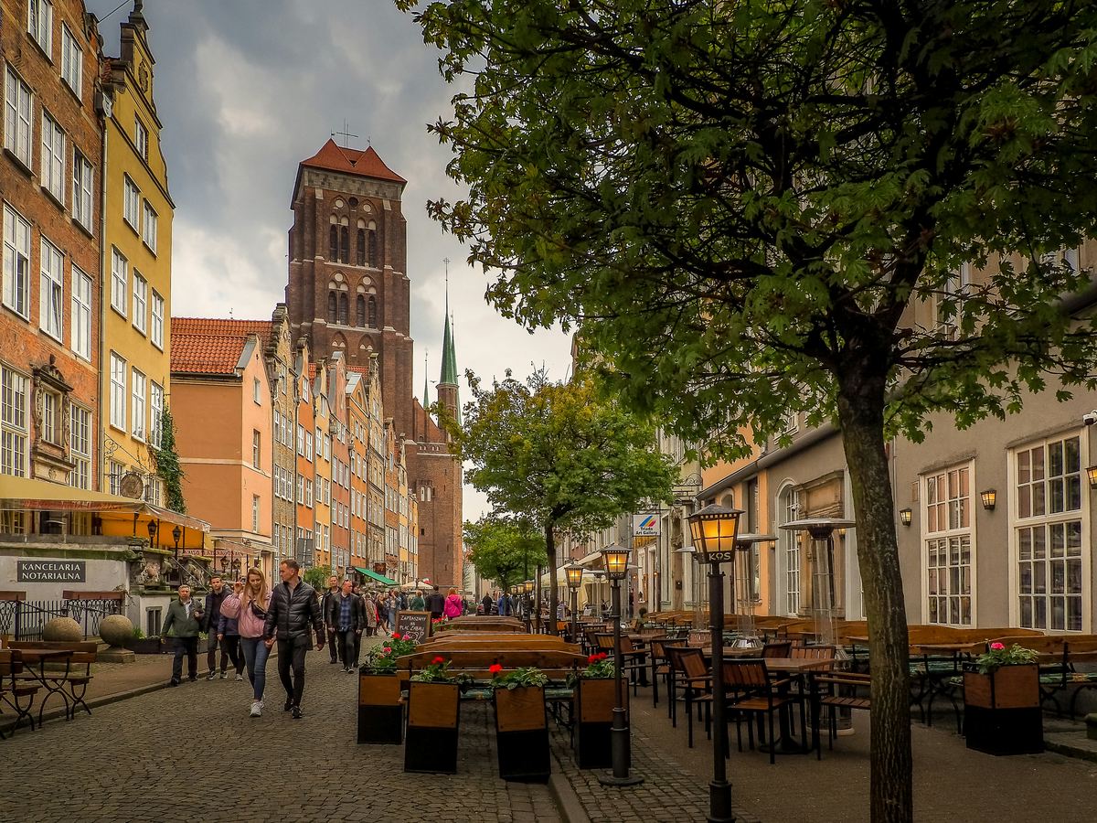 A street in Gdańsk's historic centre lined with colourful townhouses and café terraces, with the brick tower of St. Mary's Church behind
