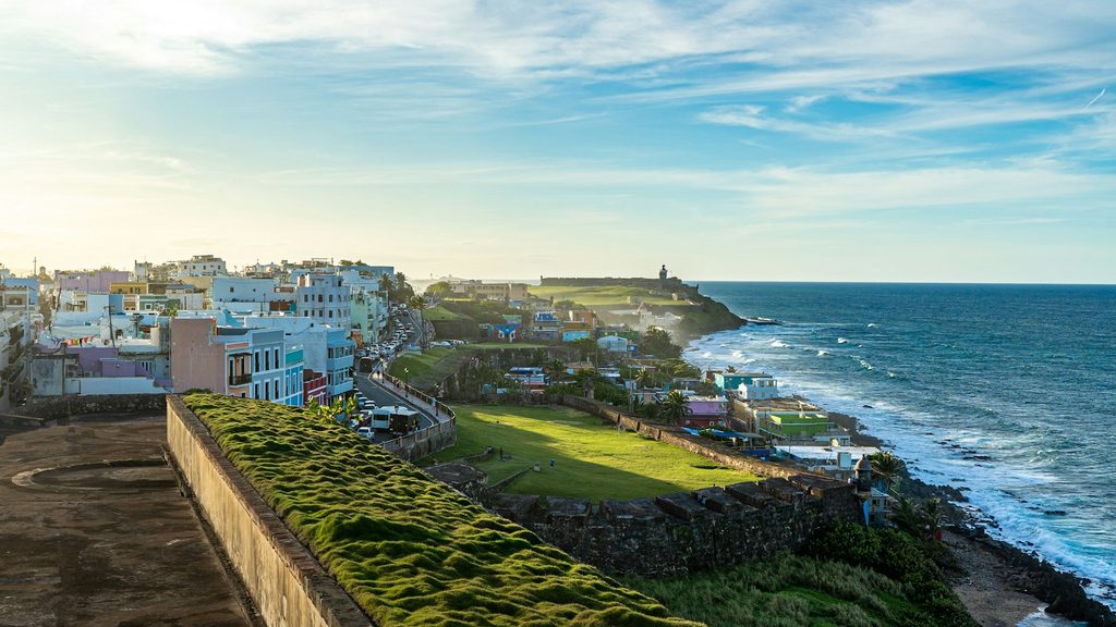 San Juan, Puerto Rico, bei Sonnenuntergang mit bunten Gebäuden entlang der Atlantikküste, historischen Steinfestungen im Vordergrund und Wellen am Ufer
