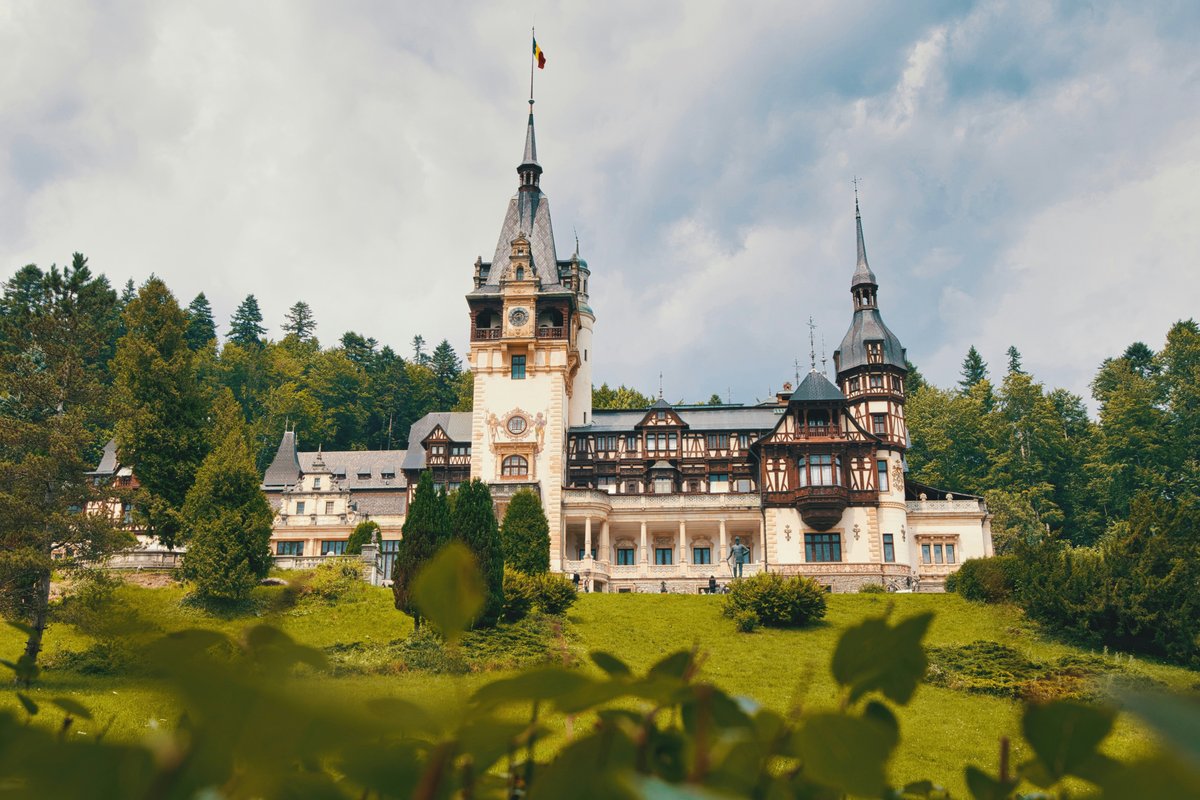 Peleș Castle's central tower and timber-framed wings above a green lawn, framed by leaves in the foreground