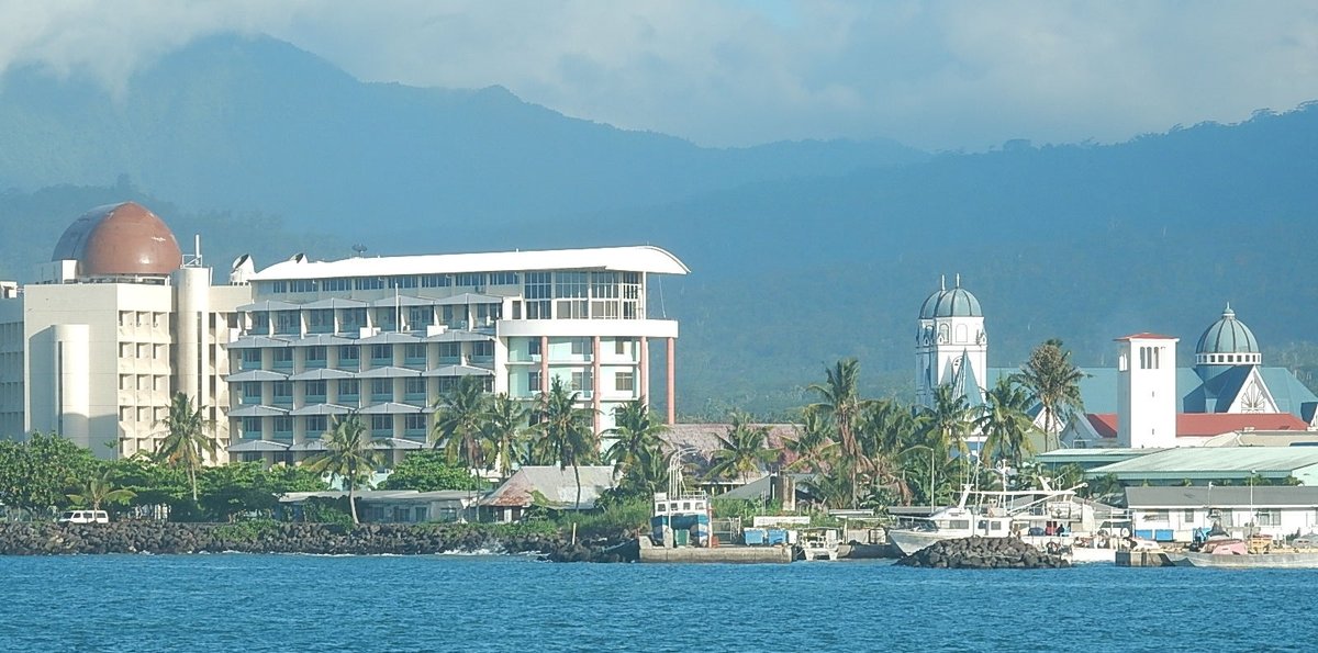 Apia Harbour in Samoa, with waterfront buildings, church towers and palm trees backed by lush volcanic mountains