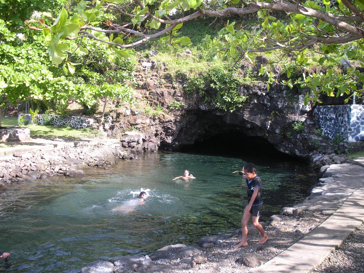 Swimmers in the clear water of Piula Cave Pool beneath a rocky cave mouth and tropical trees on Upolu