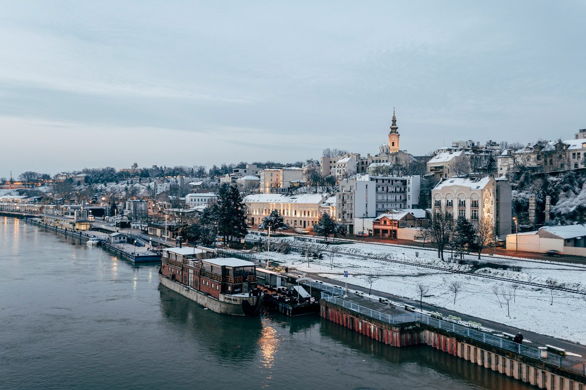 Belgrade viewed along the Sava River on a snowy winter evening, with riverside buildings, boats and the historic cityscape rising along the waterfront