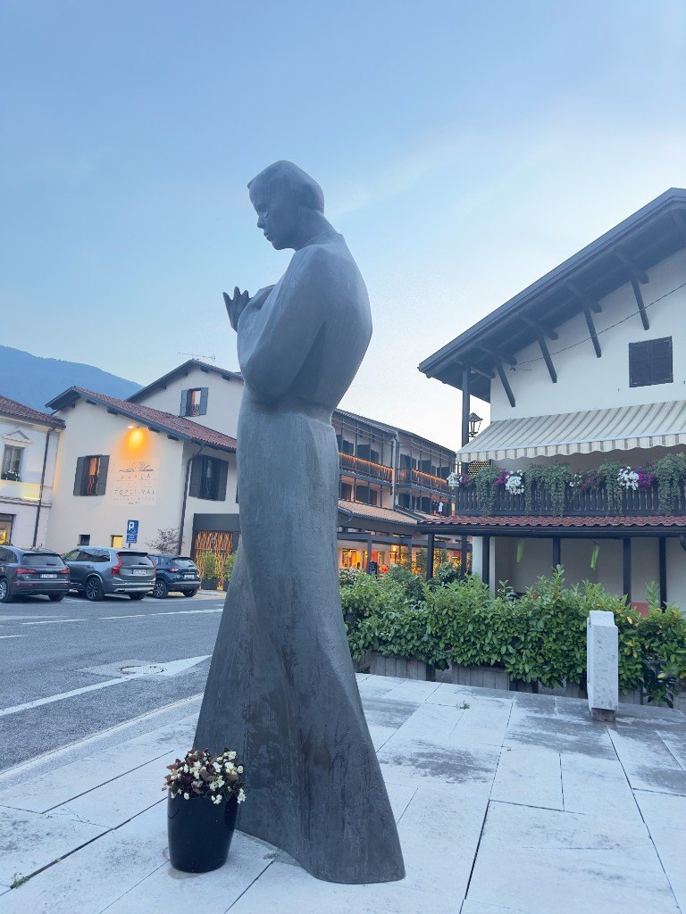 Bronze statue of a woman in Kobarid's town square, with alpine houses and evening light